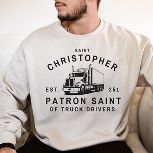 Könnte beinhalten: Sandfarbenes Sweatshirt mit schwarzer Grafik eines Sattelschleppers und dem Text "SAINT CHRISTOPHER PATRON SAINT OF TRUCK DRIVERS". Das Sweatshirt hat einen Rundhalsausschnitt und lange Ärmel.