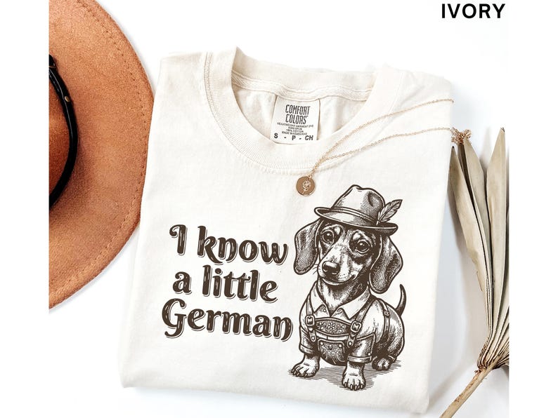 Puede incluir: Una camiseta de color marfil con un gr&aacute;fico marr&oacute;n de un perro salchicha que lleva un sombrero y pantalones de cuero. El texto "I know a little German" est&aacute; impreso en letras marrones a la izquierda del perro. La camiseta tiene una etiqueta.