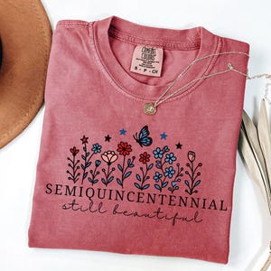 May include: A dusty rose Comfort Colors t-shirt with a floral design and the words "SEMIQUINCENTENNIAL still beautiful." A silver necklace with a pendant and a brown hat are also visible.