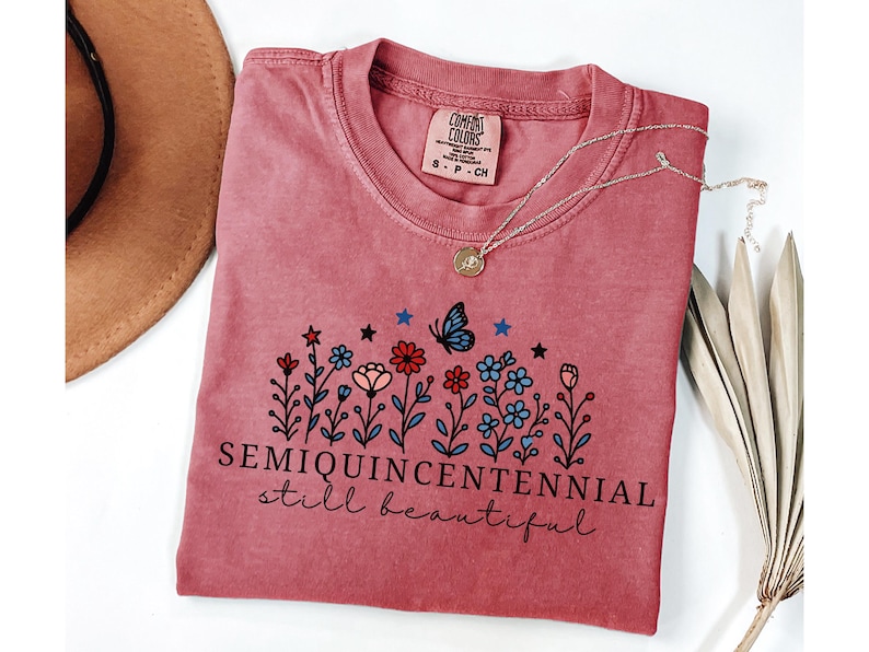 May include: A dusty rose Comfort Colors t-shirt with a floral design and the words "SEMIQUINCENTENNIAL still beautiful." A silver necklace with a pendant and a brown hat are also visible.