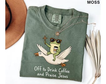 Pode incluir: Uma t-shirt verde musgo com um gráfico de um sapo usando óculos de sol com estampa de leopardo, segurando uma cruz de madeira e um copo de café enquanto monta um ganso branco voador. O texto abaixo diz Off to Drink Coffee and Praise Jesus.