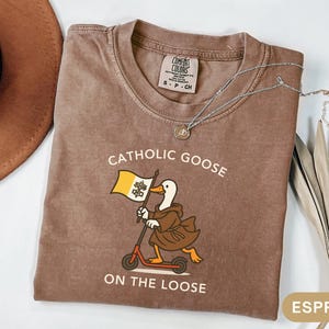 May include: Espresso-colored t-shirt with the text "Catholic Goose On The Loose" and a cartoon goose riding a scooter while holding a flag. A silver necklace with a pendant is on the shirt. The shirt is folded on a white surface.