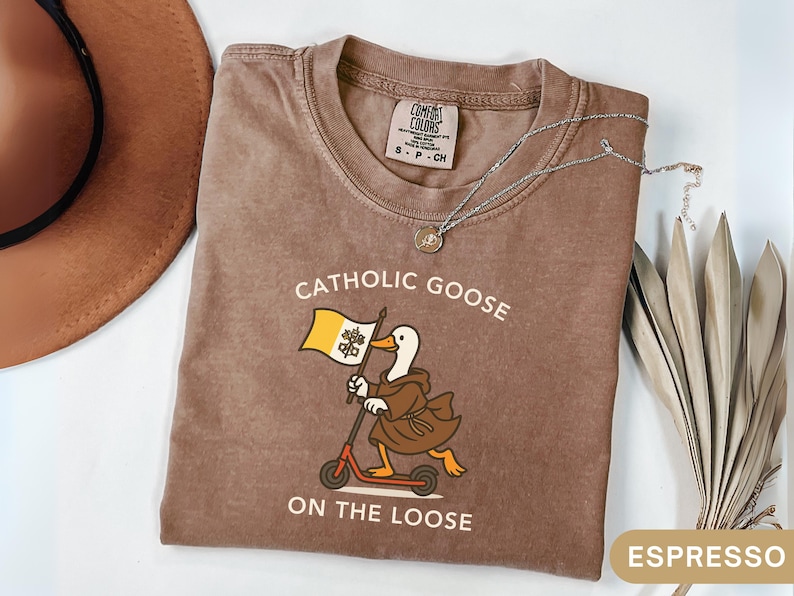 May include: Espresso-colored t-shirt with the text "Catholic Goose On The Loose" and a cartoon goose riding a scooter while holding a flag. A silver necklace with a pendant is on the shirt. The shirt is folded on a white surface.