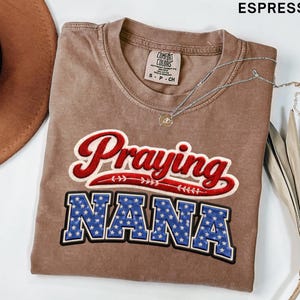 Puede incluir: Camiseta color espresso con las palabras "Praying NANA" en un diseño rojo, blanco y azul. La camiseta tiene cuello redondo y mangas cortas. Un collar plateado con un pequeño colgante está sobre la camiseta.