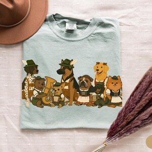 May include: A light blue t-shirt featuring a group of cartoon dogs dressed in traditional Bavarian outfits. The dogs include a Dalmatian, German Shepherd, Bulldog, Golden Retriever, and Pomeranian. Some dogs are playing instruments.