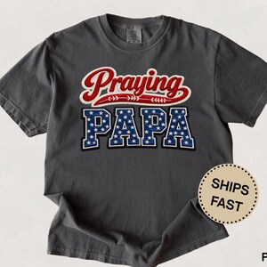 Puede incluir: Camiseta gris oscuro con las palabras "Praying PAPA" en un diseño patriótico. La palabra "Praying" está en escritura roja con detalles blancos, y "PAPA" en azul con estrellas blancas.