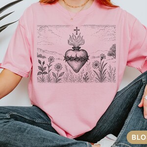 K&ouml;nnte beinhalten: Hellrosa T-Shirt mit einer schwarz-wei&szlig;en Grafik eines Herzens mit Flammen, einer Dornenkrone und einem Kreuz. Das Design ist vor einer Landschaft mit Blumen und Bergen platziert.