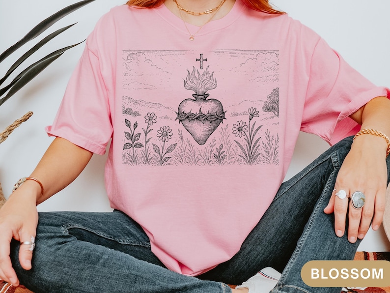 K&ouml;nnte beinhalten: Hellrosa T-Shirt mit einer schwarz-wei&szlig;en Grafik eines Herzens mit Flammen, einer Dornenkrone und einem Kreuz. Das Design ist vor einer Landschaft mit Blumen und Bergen platziert.