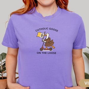 May include: Violet t-shirt with a graphic of a goose in a brown robe riding a scooter and holding a flag. The text on the shirt reads "Catholic Goose On The Loose".