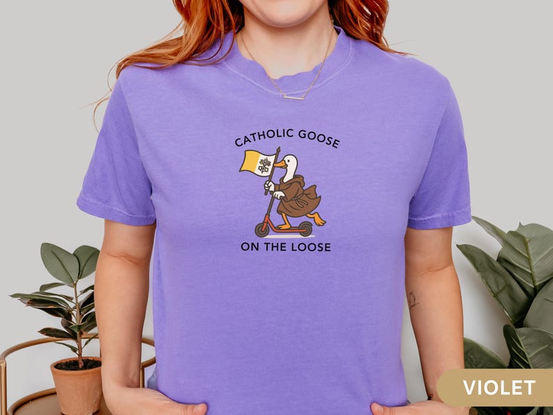 May include: Violet t-shirt with a graphic of a goose in a brown robe riding a scooter and holding a flag. The text on the shirt reads "Catholic Goose On The Loose".