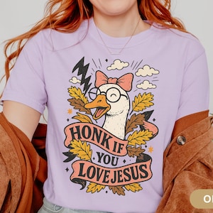 Puede incluir: Camiseta de color lila con un dibujo de un ganso con gafas y un lazo, con el texto "HONK IF YOU LOVE JESUS" en una pancarta. El dise&ntilde;o incluye rayos, nubes y hojas de oto&ntilde;o.