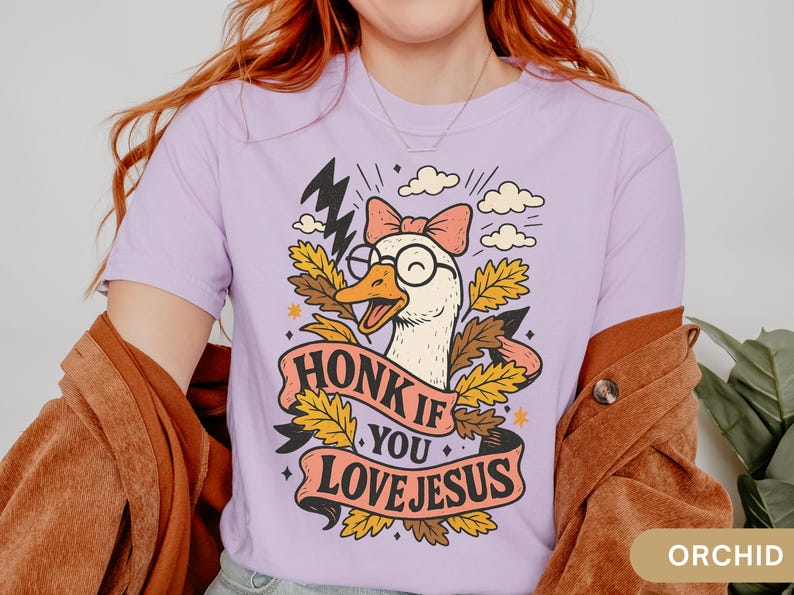 Puede incluir: Camiseta de color lila con un dibujo de un ganso con gafas y un lazo, con el texto "HONK IF YOU LOVE JESUS" en una pancarta. El dise&ntilde;o incluye rayos, nubes y hojas de oto&ntilde;o.
