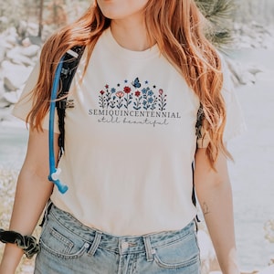 May include: Ivory-colored t-shirt with a floral design featuring red, blue, and white flowers, a butterfly, and the text "SEMIQUINCENTENNIAL still beautiful." The shirt is worn by a person in blue jeans.