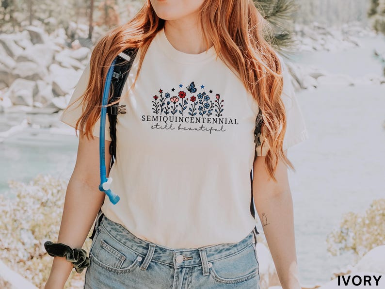May include: Ivory-colored t-shirt with a floral design featuring red, blue, and white flowers, a butterfly, and the text "SEMIQUINCENTENNIAL still beautiful." The shirt is worn by a person in blue jeans.