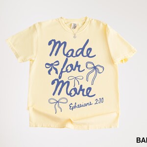 Puede incluir: Camiseta amarilla clara con la frase azul "Made for More", adornada con lazos y la referencia "Efesios 2:10". La camiseta, con cuello redondo y mangas cortas, es ideal para un look casual.