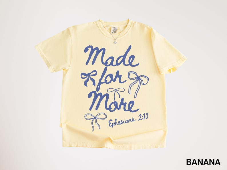 Puede incluir: Camiseta amarilla clara con la frase azul "Made for More", adornada con lazos y la referencia "Efesios 2:10". La camiseta, con cuello redondo y mangas cortas, es ideal para un look casual.