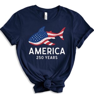 Puede incluir: Camiseta azul marino con un dise&ntilde;o de tibur&oacute;n que incorpora la bandera estadounidense. El texto dice "AMERICA" y "250 YEARS" en blanco. Una camiseta de tem&aacute;tica patri&oacute;tica.
