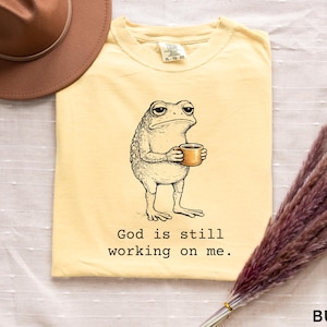 May include: Butter-colored t-shirt featuring a cartoon frog holding a coffee cup. The frog has a weary expression. The text "God is still working on me" is printed below the frog. A brown hat is in the upper left corner.