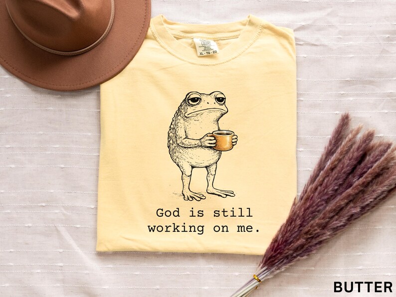 May include: Butter-colored t-shirt featuring a cartoon frog holding a coffee cup. The frog has a weary expression. The text "God is still working on me" is printed below the frog. A brown hat is in the upper left corner.