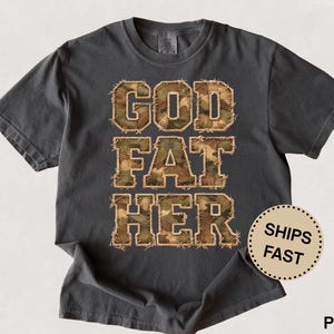 May include: Dark gray t-shirt with the words "GOD FAT HER" in a camouflage pattern. The shirt has a round neck and short sleeves. A circular sticker with the words "SHIPS FAST" is on the bottom right.