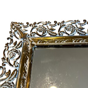 Pode incluir: Close-up de um espelho decorativo com uma moldura dourada ornamentada. A moldura apresenta uma sobreposi&ccedil;&atilde;o branca com desenhos florais e de rolagem. A superf&iacute;cie do espelho &eacute; reflexiva, e a est&eacute;tica geral &eacute; vintage.