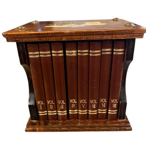 May include: A wooden book holder with eight volumes. The books are brown with gold lettering and trim. The holder is dark brown with decorative gold accents on the top and corners. The volumes are labeled I through VIII.