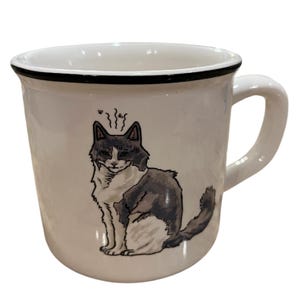 May include: White ceramic mug with a black rim and handle. The mug features a whimsical illustration of a gray and white cat with steam rising from its head. The cat is sitting, and the mug has a glossy finish.