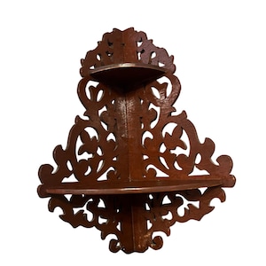 Può includere: Mensola angolare in legno marrone scuro a due livelli. La mensola presenta intricati disegni floreali e a volute intagliati. I bordi delle mensole sono arrotondati. La mensola è progettata per adattarsi a un angolo.