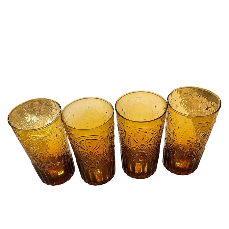 May include: Four amber-colored drinking glasses. Each glass has a textured, embossed design around the body. The glasses are cylindrical with a slightly flared top. The design appears to be a repeating pattern of floral or geometric shapes.