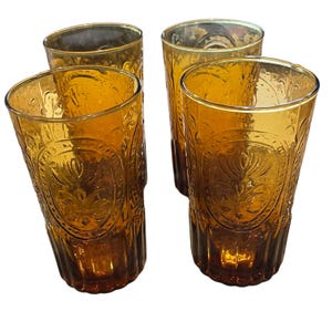 May include: Four amber-colored drinking glasses. Each glass has a tall, cylindrical shape with a decorative embossed pattern. The glasses are made of a transparent material, allowing light to pass through, and are suitable for serving beverages.