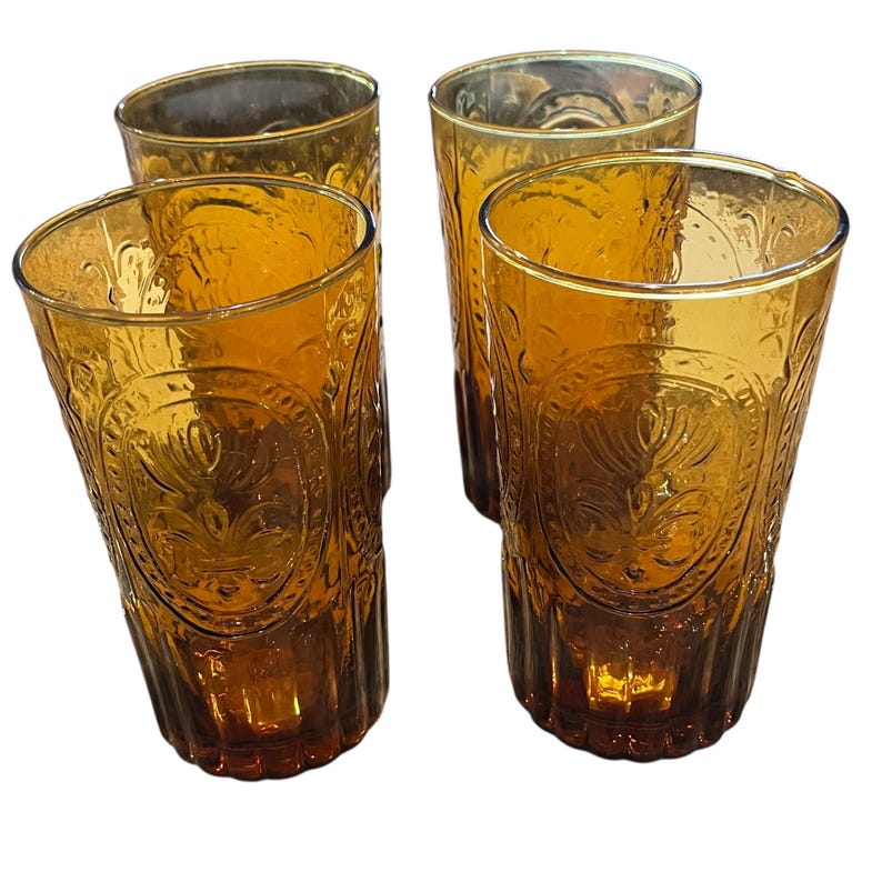 May include: Four amber-colored drinking glasses. Each glass has a tall, cylindrical shape with a decorative embossed pattern. The glasses are made of a transparent material, allowing light to pass through, and are suitable for serving beverages.