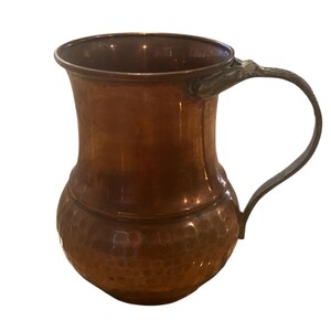 Puede incluir: Una jarra de cobre con una textura martillada en la mitad inferior y una parte superior lisa. El asa es de un metal más oscuro, y la jarra tiene una base bulbosa y un borde acampanado. La jarra es probablemente vintage.