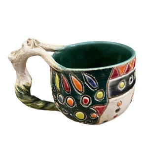 May include: A handcrafted ceramic mug with a unique, textured handle. The mug features a dark green interior and exterior with colourful, abstract designs in red, yellow, orange, and blue. The handle has a branch-like appearance.