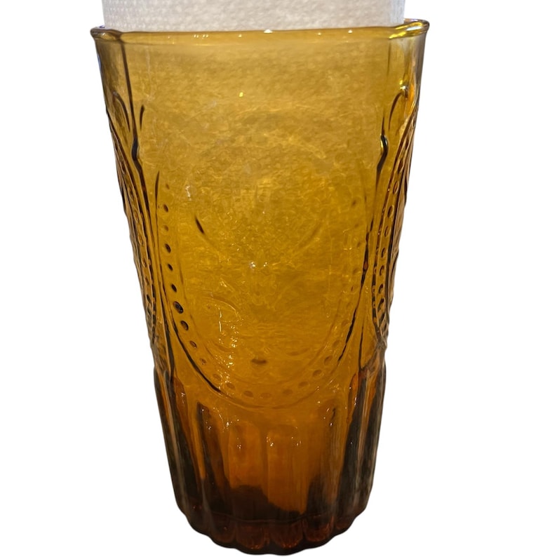 May include: Amber-colored glass tumbler with embossed floral and dot patterns. The tall, cylindrical glass has a slightly flared top and a thick base. The glass is empty and sits against a white background.