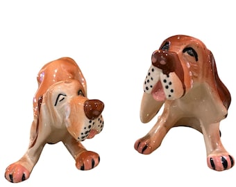 May include: Two ceramic dog figurines. One is in a playful pose, the other is sitting up. Both dogs have brown and tan fur with black spots and pink accents on their paws and ears. The figurines are on a white background.