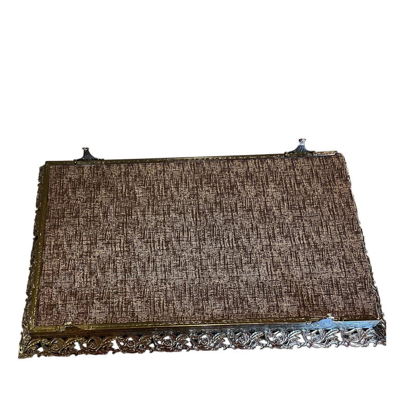 Pode incluir: Bandeja de vaidade retangular vintage com uma inser&ccedil;&atilde;o de tecido castanho e bege texturizado. A bandeja tem uma moldura de metal dourado decorativa com detalhes ornamentados ao longo das bordas e pequenos detalhes decorativos nos cantos.
