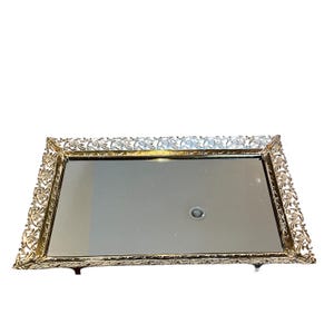 Pode incluir: Uma bandeja retangular espelhada com uma borda de filigrana dourada ornamentada. A bandeja tem uma superf&iacute;cie reflexiva e &eacute; suportada por pequenos p&eacute;s. A borda decorativa apresenta desenhos florais e de rolagem.