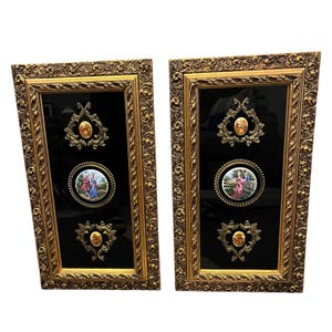 May include: Two rectangular wall plaques with ornate gold-colored frames and black backgrounds. Each plaque features a central circular porcelain medallion depicting figures in period clothing, flanked by two smaller oval porcelain decorative accents.