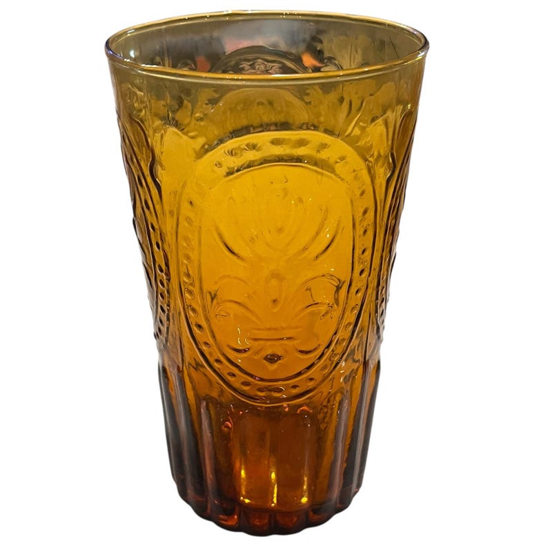 May include: Amber-colored drinking glass with an embossed fleur-de-lis design. The tall glass has a textured pattern and a wide rim. The glass is made of a transparent material and is suitable for beverages.