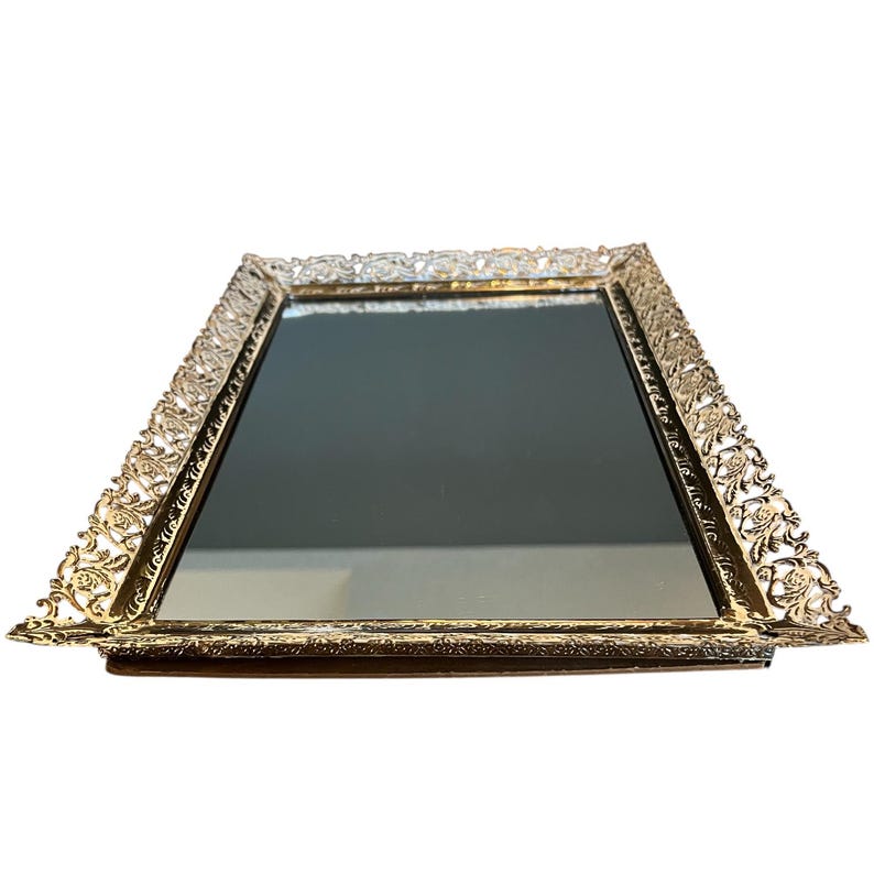 Pode incluir: Uma bandeja de toucador retangular com uma superf&iacute;cie espelhada e uma moldura de filigrana dourada ornamentada. A moldura apresenta um design floral e de rolagem detalhado. A bandeja &eacute; provavelmente feita de metal e &eacute; adequada para exibir perfumes ou joias.