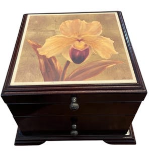 Puede incluir: Una caja de joyería de madera marrón oscuro con dos cajones y una tapa con bisagras. La tapa presenta una pintura de una orquídea amarilla y púrpura sobre un fondo dorado. Los cajones tienen pomos decorativos plateados.