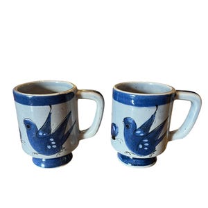 May include: Two ceramic mugs with a blue and white bird design. The mugs have a light blue base color with a dark blue rim and handle. Each mug features a blue bird illustration.
