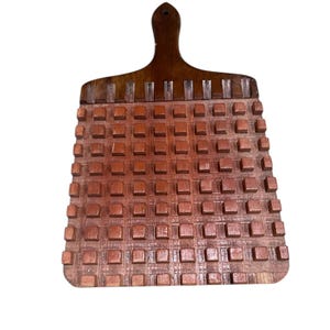 Puede incluir: Una tabla de cortar de madera vintage con asa. La tabla es de color marrón cálido y presenta un patrón de cuadrícula de cuadrados en relieve en su superficie. El asa es de un tono más oscuro y tiene un agujero para colgar.