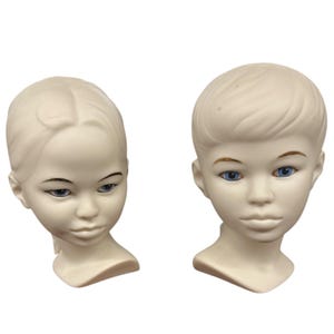 May include: Two off-white ceramic mannequin heads with blue eyes. One has a side-parted hairstyle, the other has a short, layered style. Both have painted eyebrows and lips, and are set against a white background.
