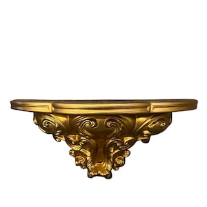 May include: A wall-mounted shelf featuring a metallic gold finish and a semi-circular top surface. The underside of the shelf is decorated with intricate, raised scrollwork and ornate carvings that taper down to a central point.