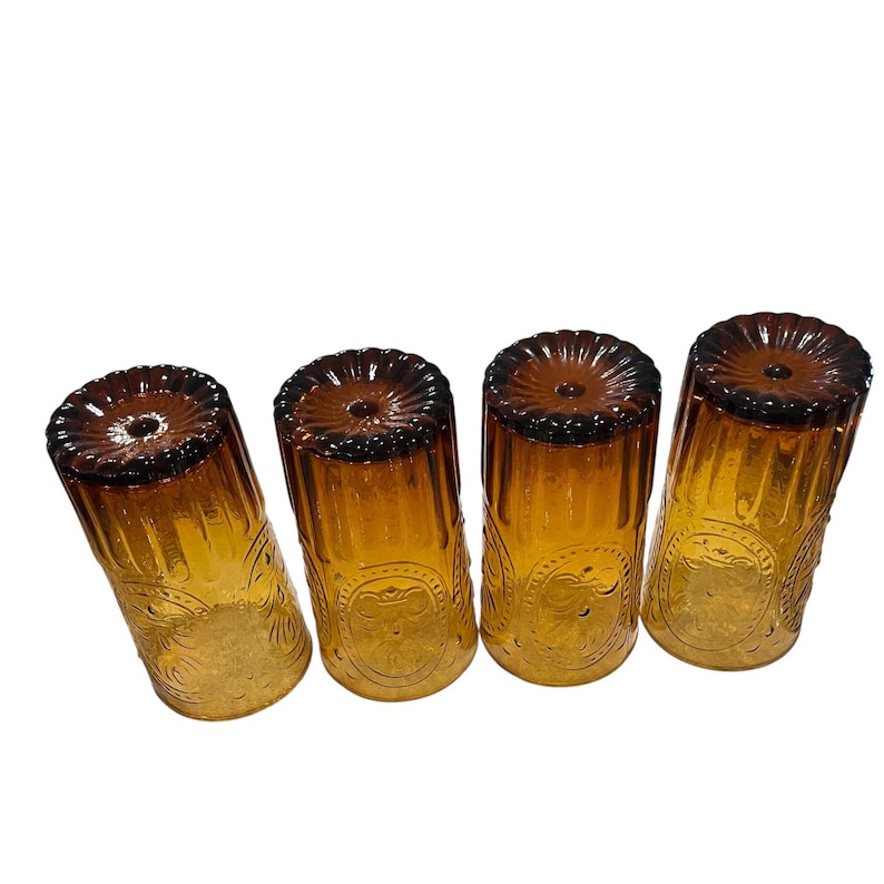 May include: Four amber-colored drinking glasses with a decorative floral pattern. The glasses have a ribbed design and a dark brown rim. The glasses are made of glass and are suitable for serving beverages.