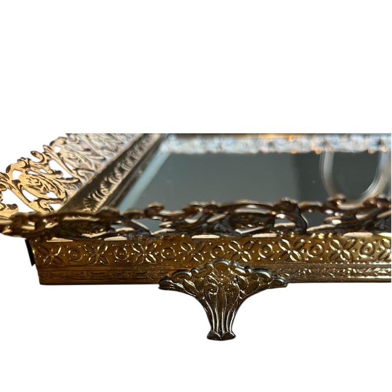 Pode incluir: Uma bandeja de vaidade dourada e ornamentada com uma superf&iacute;cie espelhada. A bandeja apresenta detalhes intrincados de flores e rolagem nas bordas e na base. Uma al&ccedil;a decorativa &eacute; vis&iacute;vel na frente. A bandeja &eacute; provavelmente vintage.