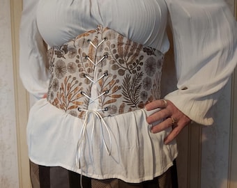 Könnte beinhalten: Eine weiße Bluse mit langen Ärmeln und einem braunen und goldenen Korsett-Gürtel mit Blumenmuster.