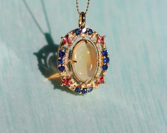 May include: A gold pendant necklace featuring an oval opal gemstone, surrounded by a halo of blue and pink gemstones. The pendant hangs from a delicate gold chain. The text "AFRA JEWELRY STUDIO" is at the bottom.