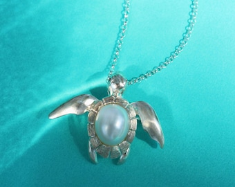 May include: A silver-toned turtle pendant necklace with a pearl center, set against a teal background. The pendant features detailed flipper and head designs. The necklace is from AFRA JEWELRY STUDIO.